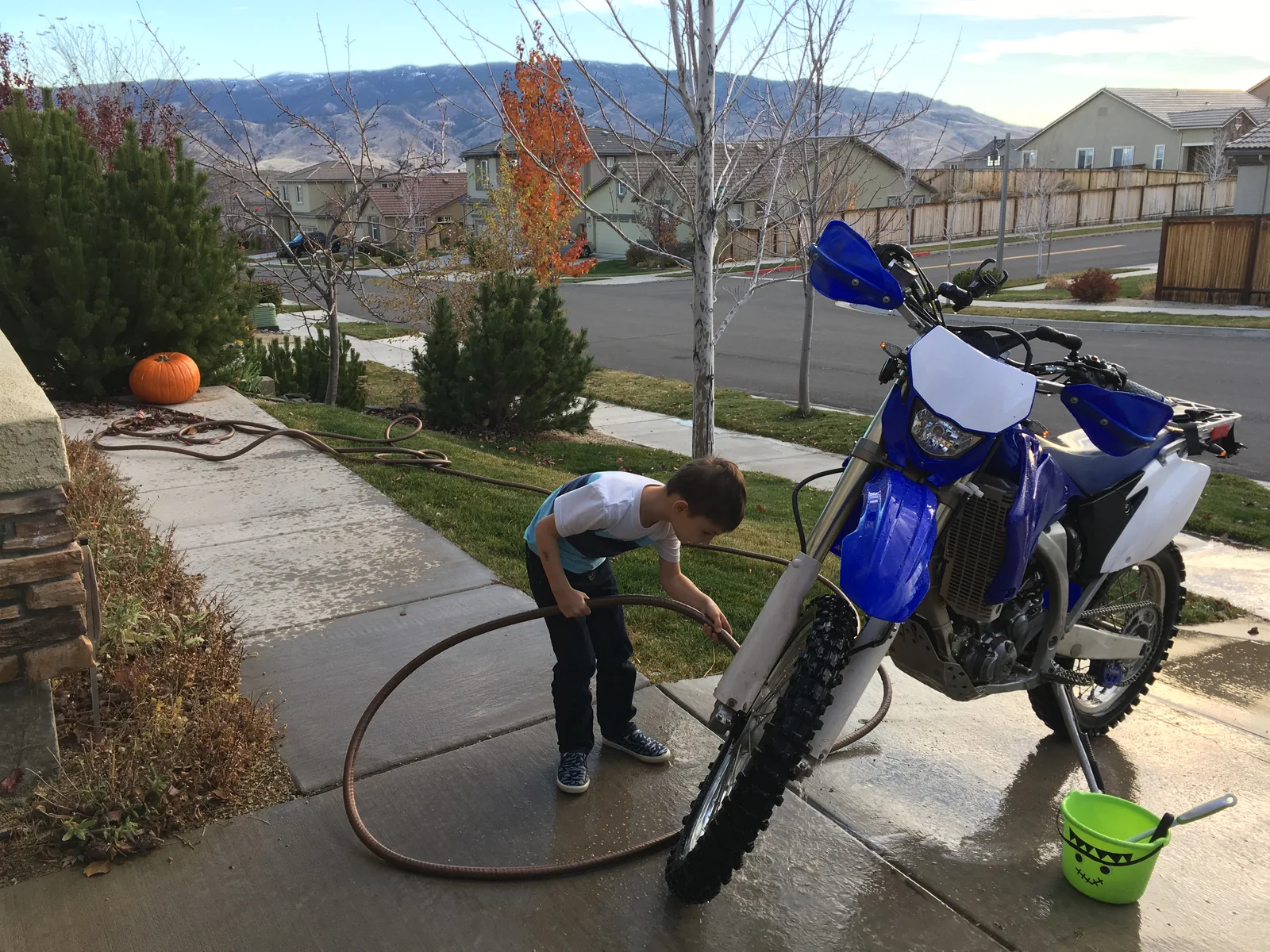 Blue dirt bike being washed in a driveway while a child sprays the front wheel with a hose.