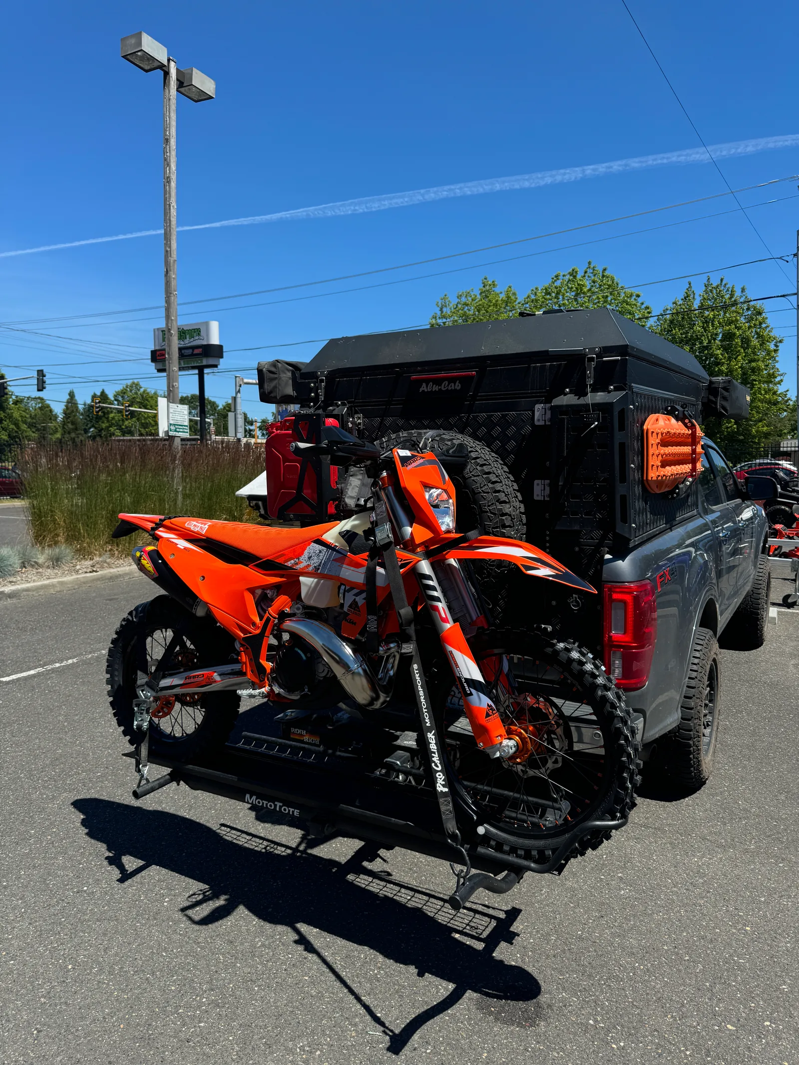 Orange KTM dirt bike secured to the back of a pickup truck on a sunny day.
